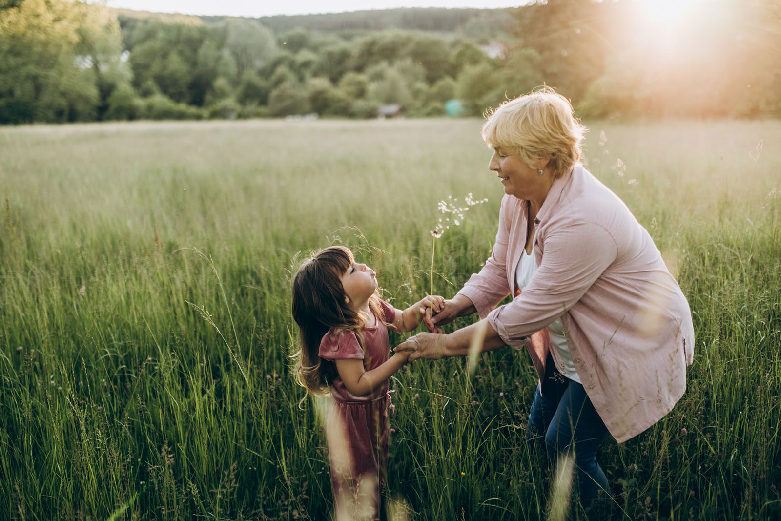 An older woman and a young girl hold hands and smile at each other while standing in a grassy field at sunset.