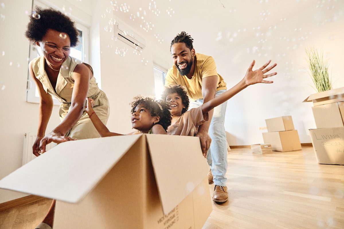 A family of four smiles and plays with cardboard boxes in a bright room with moving boxes