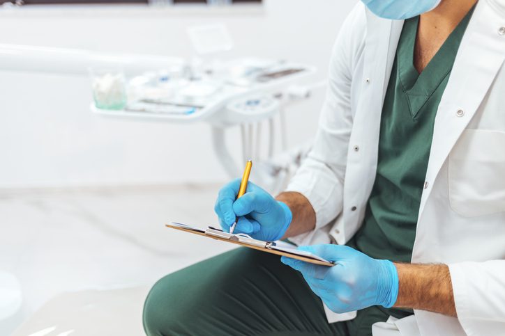 A healthcare professional in scrubs and gloves writes on a clipboard in a medical or dental office.