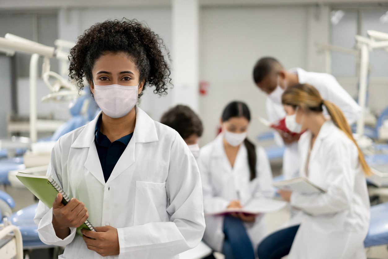 A woman in a lab coat and mask holds a notebook in a dental clinic