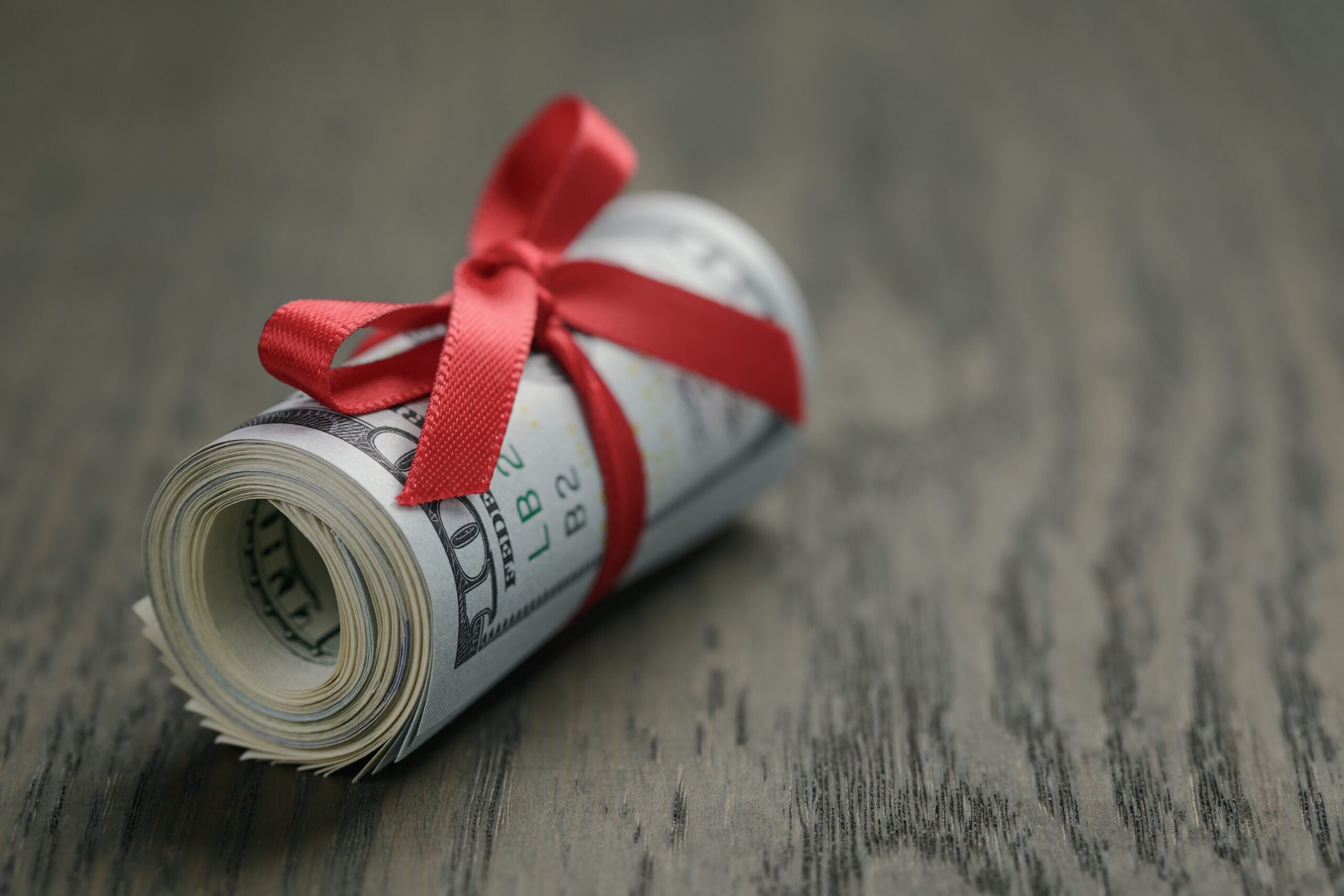 A roll of U.S. dollar bills tied with a red ribbon sits on a wooden surface.