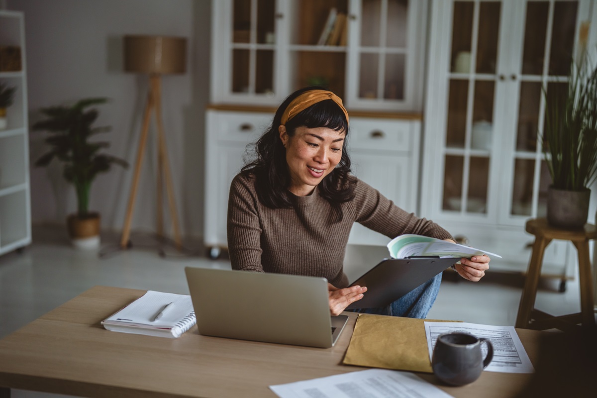 Asian woman smiling and concentrating while working remotely from her spacious home office, reviewing paperwork and using a laptop for productivity and business tasks
